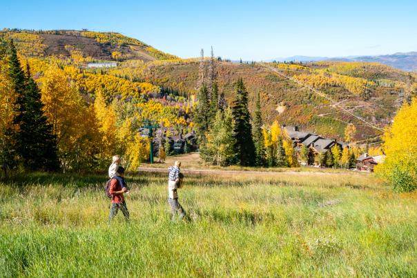 Two parents hike with their children riding on shoulders on a trail in front of Fall leaves in Park City, UT