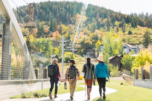 Four hikers walk across a bridge toward ski runs and fall foliage on a hillside