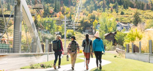 Four hikers walk across a bridge toward ski runs and fall foliage on a hillside