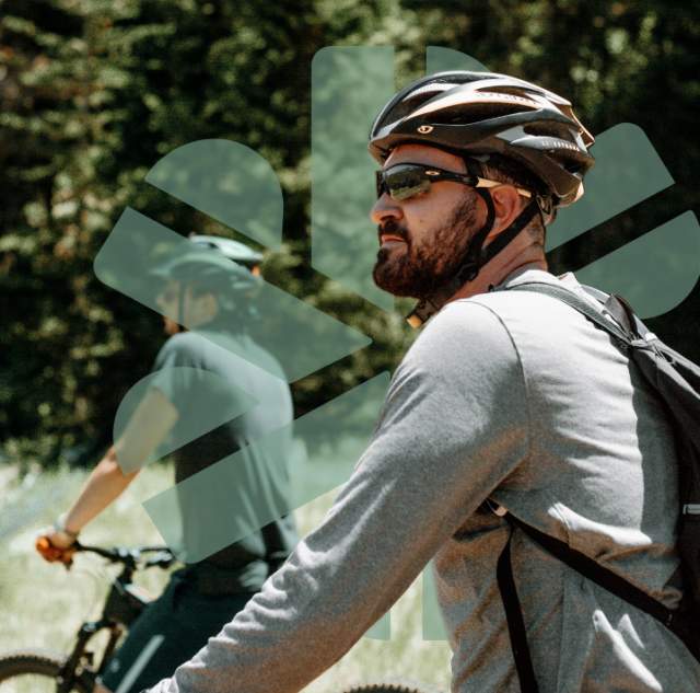 a couple of bicyclers ride outdoors; a logo is embedded behind the foreground biker