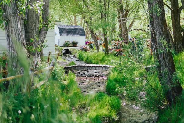 Photo from the outside of Park City Gardens. Plants and flowers in the foreground with a short trail leading to a small white barn.